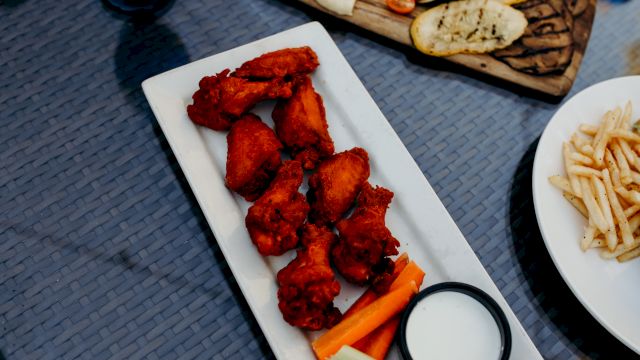 A platter of spicy red fried chicken with carrot/celery sticks and a side of white dip, plus fries and flatbread in the background.