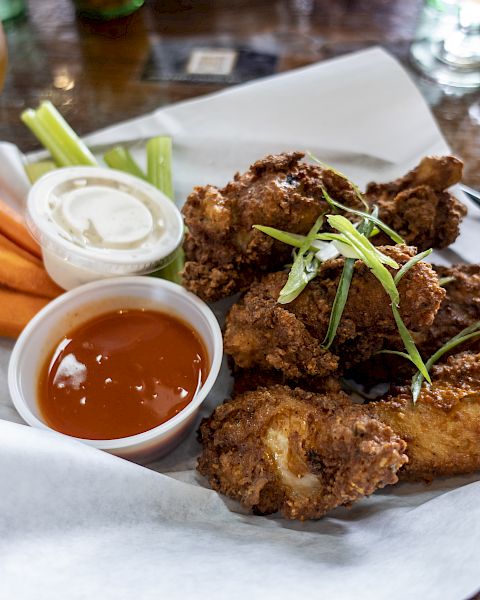 A plate of fried chicken wings with sliced green onions, accompanied by carrot sticks, celery, ranch, and hot sauce, served with a drink.
