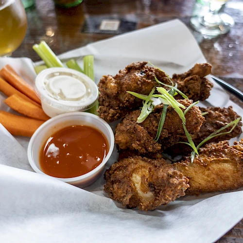 A plate of fried chicken wings with sliced green onions, accompanied by carrot sticks, celery, ranch, and hot sauce, served with a drink.