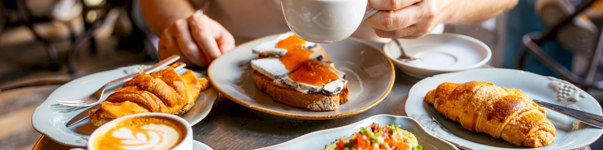 Brunch scene: a hand holds a latte over plates with toast, croissants, and orange-topped egg dishes.
