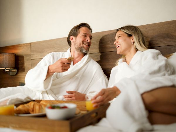 A couple in bathrobes enjoying breakfast in bed with croissants, juice, and fruit, sharing a lighthearted moment in a cozy setting.