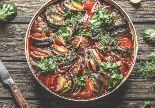 This image shows a dish of ratatouille with sliced vegetables and fresh herbs on a wooden table.