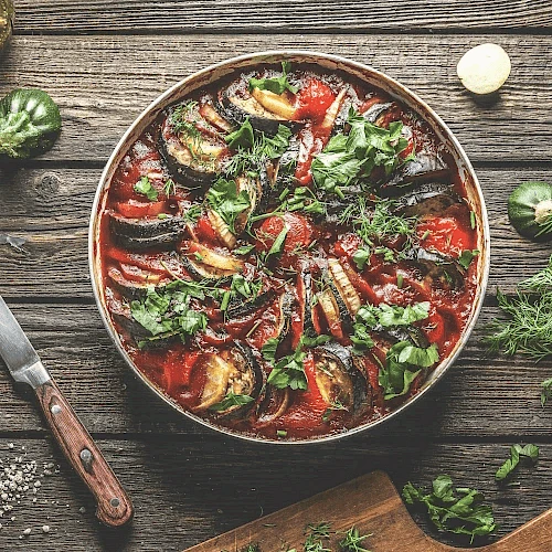 This image shows a dish of ratatouille with sliced vegetables and fresh herbs on a wooden table.