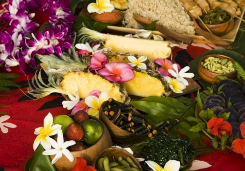 A vibrant spread of tropical fruits, colorful flowers, and assorted dishes on a red cloth, conveying a festive and exotic atmosphere.