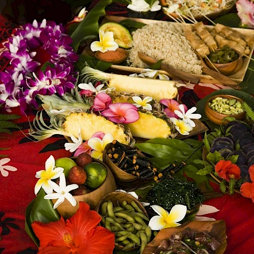 A vibrant spread of tropical fruits, colorful flowers, and assorted dishes on a red cloth, conveying a festive and exotic atmosphere.