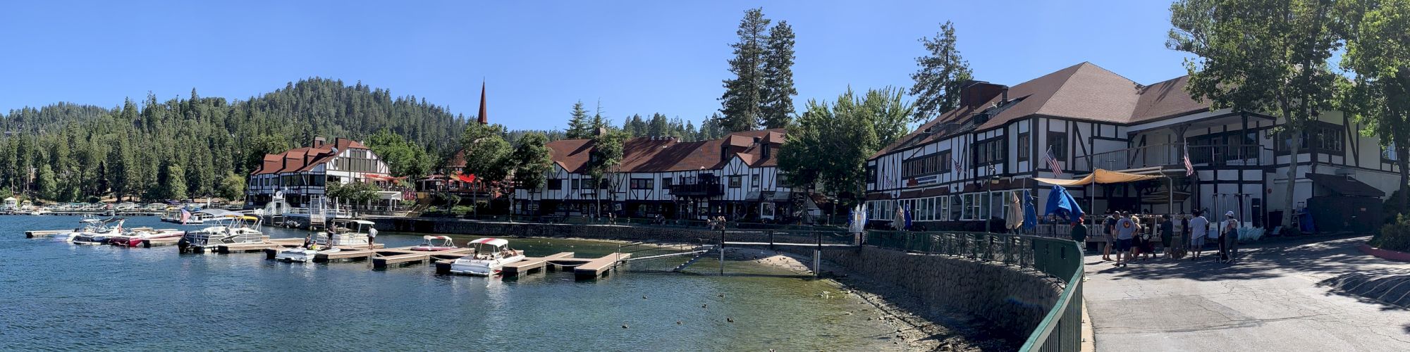A lakeside marina with many docked boats, a promenade, and Tudor-style buildings under a clear blue sky.