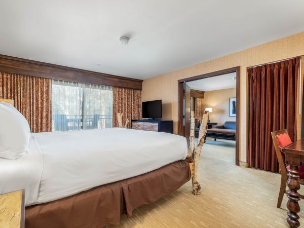 The image shows a hotel room with a bed, curtains, a desk, a TV, and an adjacent sitting area with a sofa and lamp, all in warm tones.