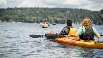 Two people in a bright orange kayak paddle across a calm lake, wearing life jackets, with forested hills in the distance.