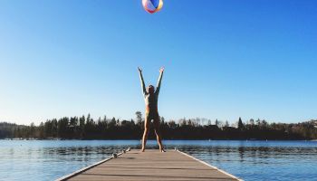 A person stands triumphantly at the end of a wooden dock on a calm lake, arms raised as a beach ball soars above under a clear blue sky.