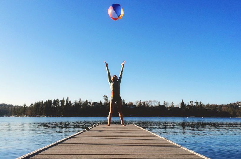 A person stands triumphantly at the end of a wooden dock on a calm lake, arms raised as a beach ball soars above under a clear blue sky.