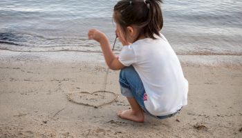 A girl sits on the beach drawing a heart in the sand near the water, wearing a white shirt and jeans, focused on her art.