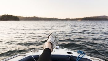 A person&rsquo;s legs and sneakers stretched out on a small boat, feet up, with a calm lake and distant shoreline at sunset.