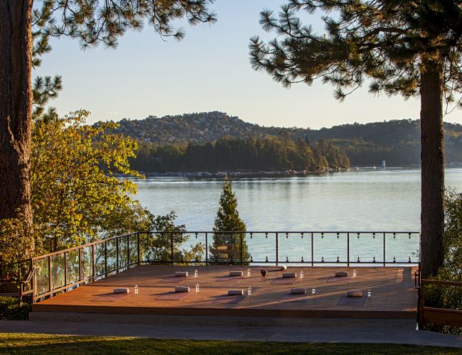 A sunny lakeside terrace with a railing, overlooking a calm lake framed by trees and distant hills.