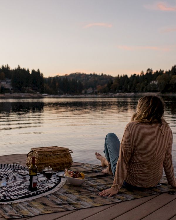 A person sits on a dock by a calm lake at sunset, with a picnic blanket, a basket, and a drum nearby, gazing at the water.