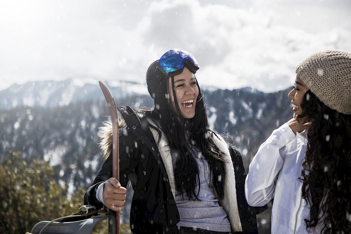 Two people smiling outdoors in snowy mountains, one holding skis, both in winter clothing, enjoying a snowy day.