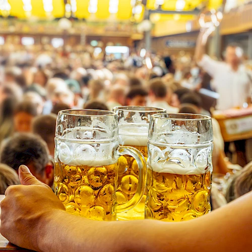 A person holds large mugs of beer at a crowded, lively festival setting, with people and colorful decorations in the background.