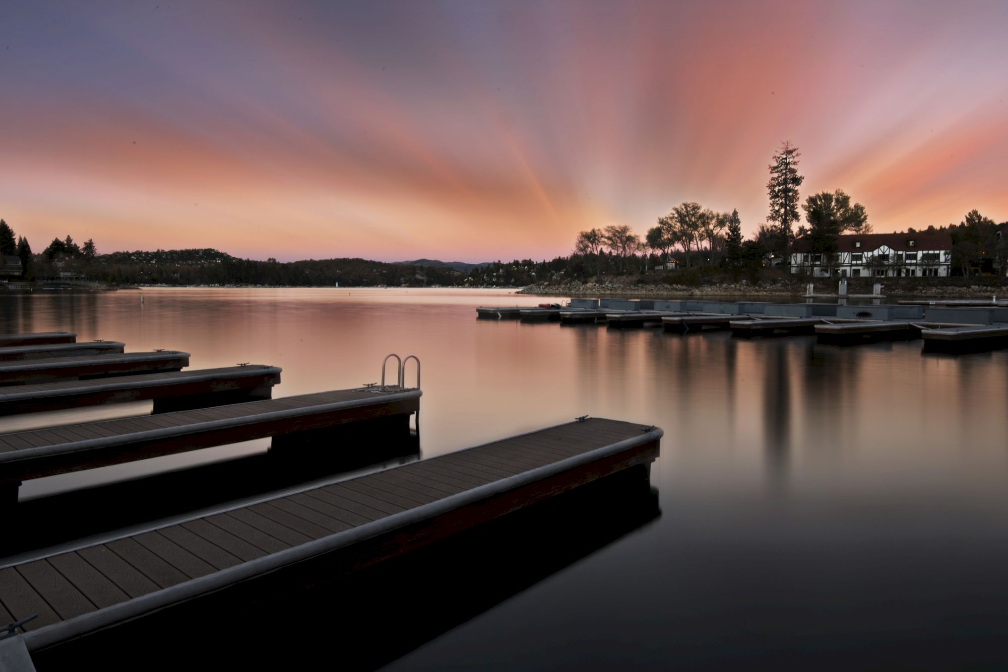 A tranquil lakeside scene at sunset with wooden docks jutting into calm water, soft pastel sky, and silhouettes of trees and distant buildings, serene.