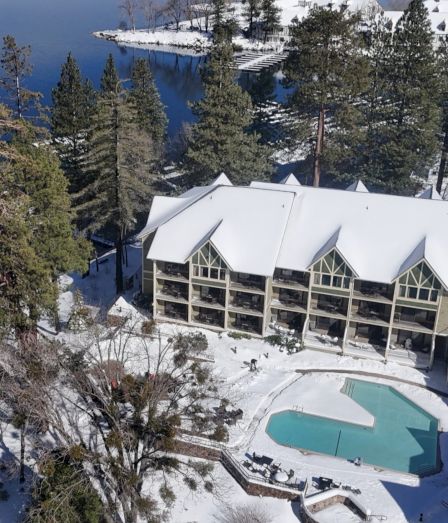 A snowy lakeside resort with white-roof buildings, an outdoor pool, pine trees, and a frozen shoreline under bright sunlight.