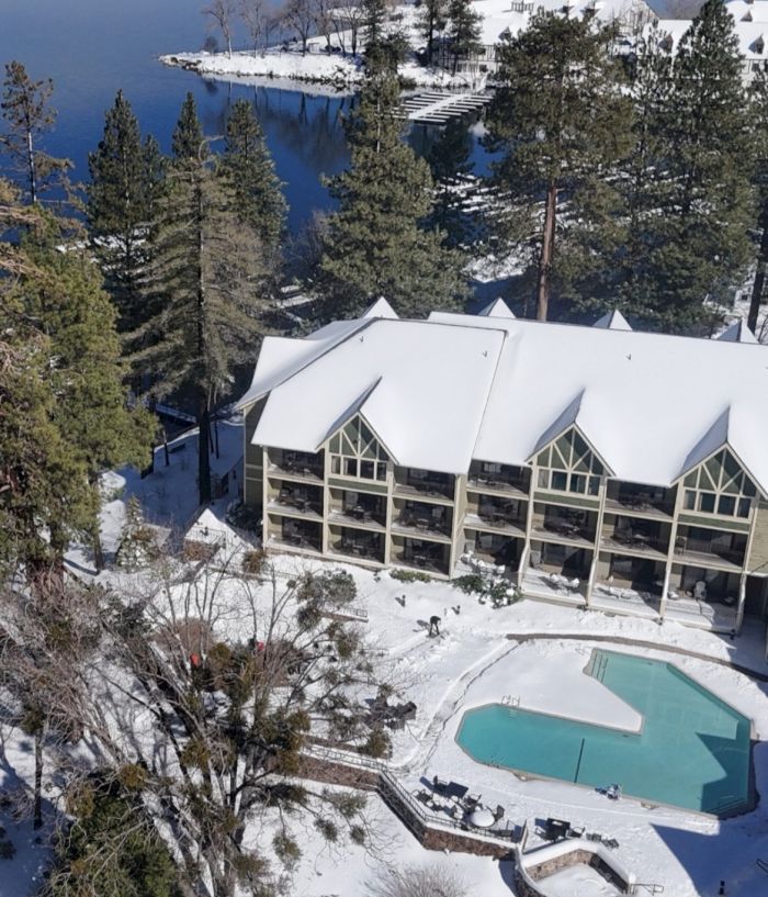 A snowy lakeside resort with white-roof buildings, an outdoor pool, pine trees, and a frozen shoreline under bright sunlight.