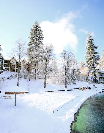 A snowy riverside resort with pine trees dusted in snow, bright blue sky, and long hotel balconies beside a green canal.