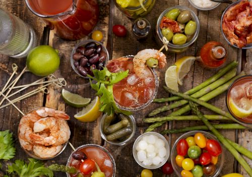The image shows a variety of appetizers and garnishes, including shrimp, olives, cherry tomatoes, and mixed beverages on a rustic wooden table.