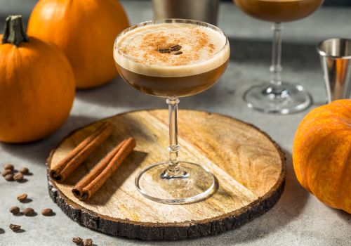 A cocktail topped with foam on a glass, cinnamon sticks, pumpkins and coffee beans on a rustic wooden slice.