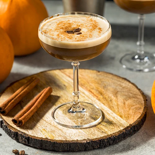 A cocktail topped with foam on a glass, cinnamon sticks, pumpkins and coffee beans on a rustic wooden slice.