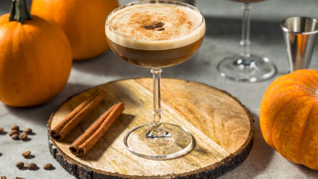 A cocktail topped with foam on a glass, cinnamon sticks, pumpkins and coffee beans on a rustic wooden slice.