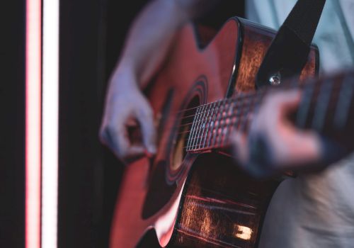 A person plays an acoustic guitar, strumming near a neon-lit edge, focus on strings and frets with a moody, intimate vibe.
