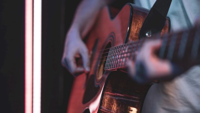 A person plays an acoustic guitar, strumming near a neon-lit edge, focus on strings and frets with a moody, intimate vibe.