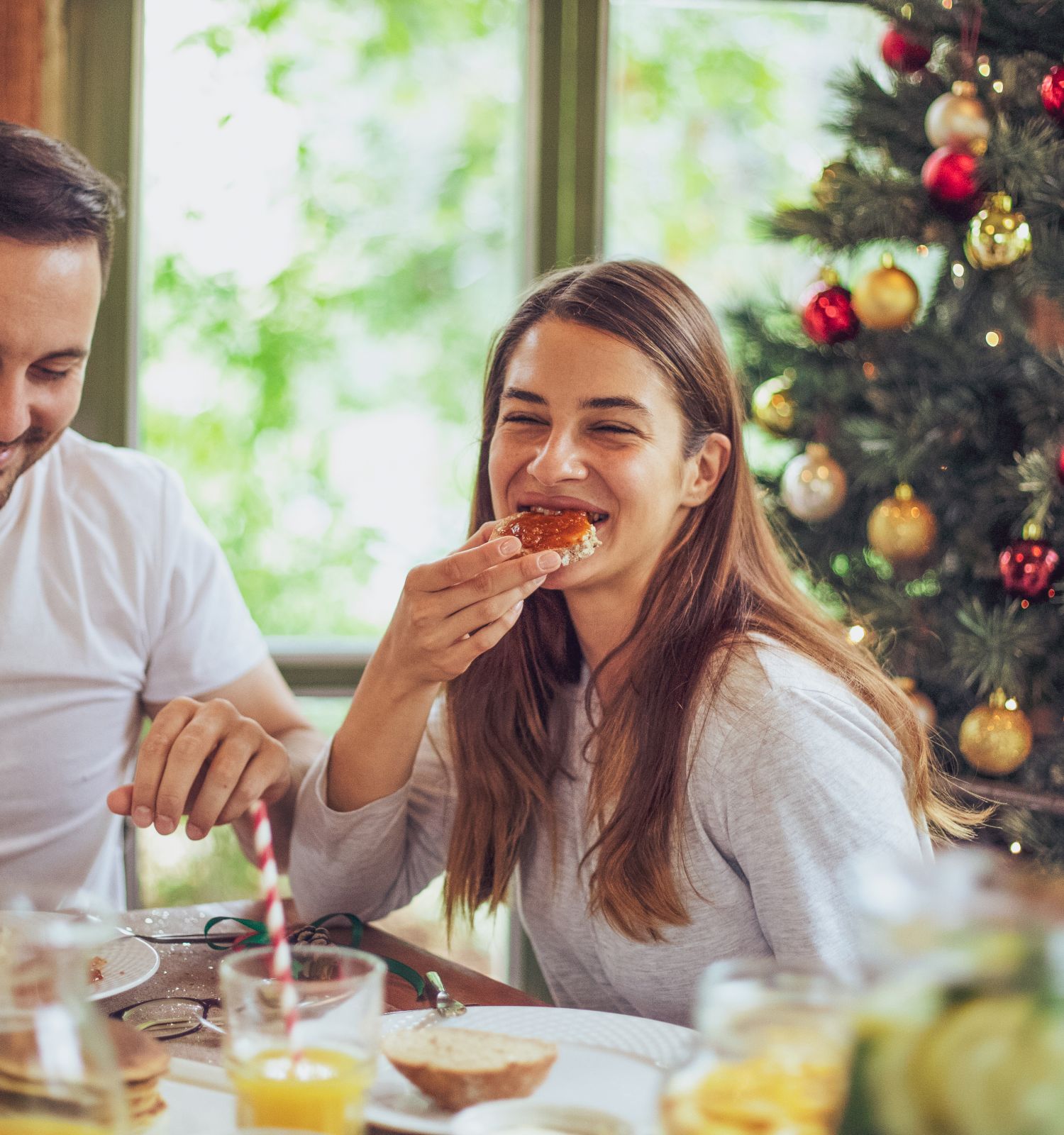 A couple enjoys a holiday breakfast near a Christmas tree, sharing pastries and juice at a cozy table. Endearing, festive morning vibes.
