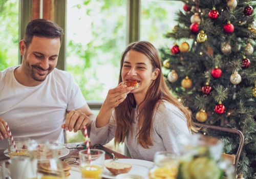 A couple enjoys a festive breakfast by a Christmas tree, sharing a bite of pastry, with bright natural light and a cheerful, cozy scene.