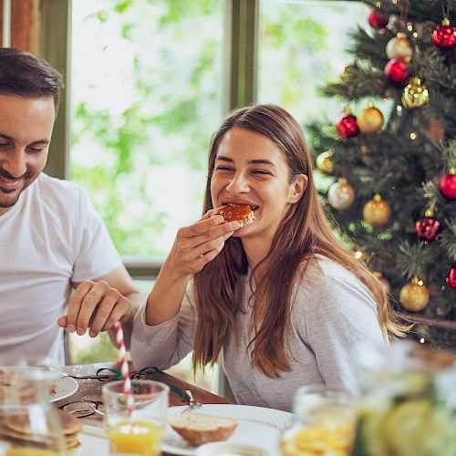 A couple enjoys a festive breakfast by a Christmas tree, sharing a bite of pastry, with bright natural light and a cheerful, cozy scene.