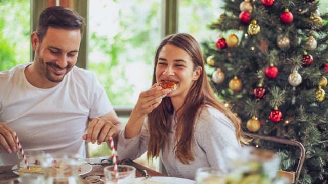 A couple enjoys a festive breakfast by a Christmas tree, sharing a bite of pastry, with bright natural light and a cheerful, cozy scene.