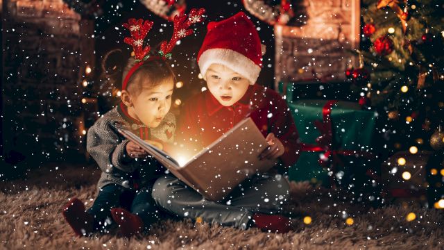 Two kids sit by a fireplace in festive hats, glowing from a shared book as snow falls around a cozy Christmas scene.