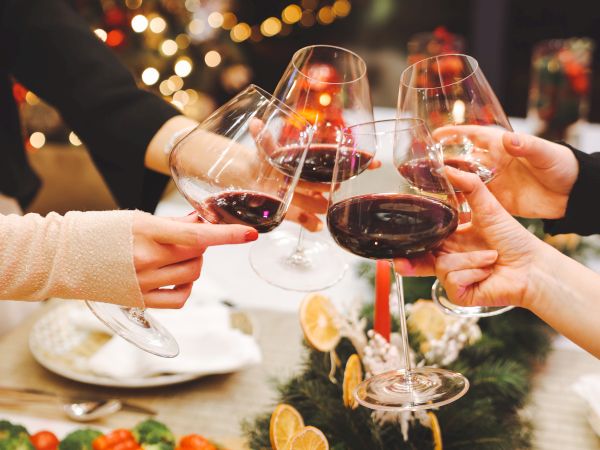 A festive toast: people raise glasses of red wine in a warmly lit holiday dinner, candles and greenery creating a cozy celebratory scene.