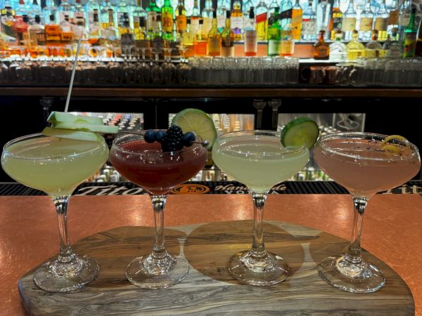 A row of four cocktail glasses on a wooden board with lime wheels, set against a colorful bar backdrop.
