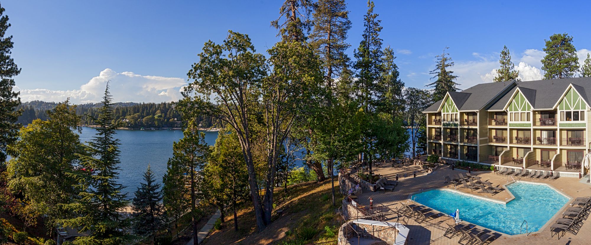 A lakeside resort with a pool, lounge chairs, and tall trees by calm water under a blue sky.