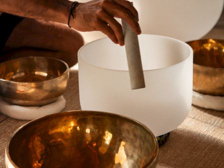 A hand holds a white mallet, playing a set of singing bowls on a textured mat, with warm golden bowls around in a calming, meditative scene.