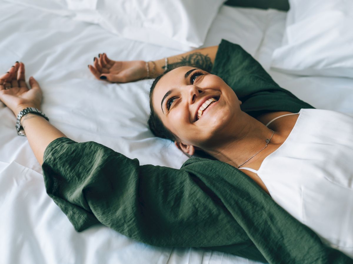 A smiling woman lies on a bed with white sheets, arms stretched out, wearing a white top and a green cardigan.