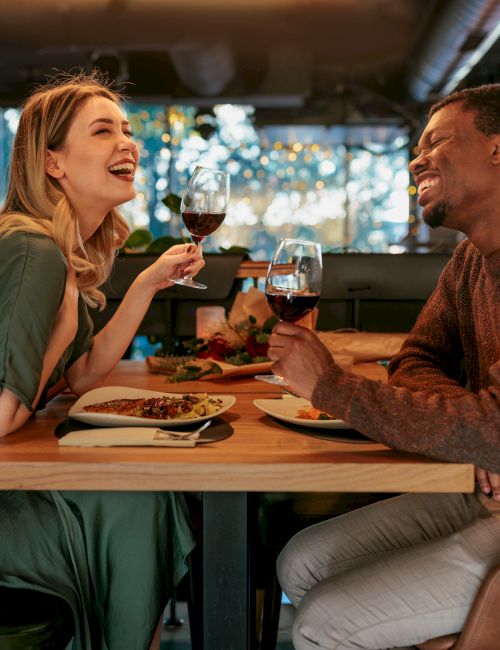 A couple enjoys a cozy dinner at a restaurant, clinking glasses and smiling across the table.