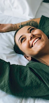 A smiling woman lies on a bed with white sheets, arms stretched out, wearing a white top and green cardigan. (140 chars)