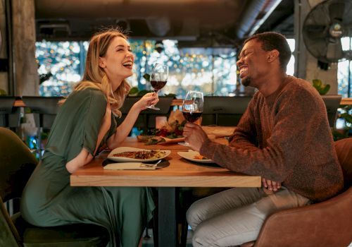 Two people share a warm dinner at a cozy restaurant, clinking glasses and laughing across a wooden table.