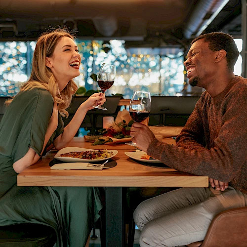 Two people share a warm dinner at a cozy restaurant, clinking glasses and laughing across a wooden table.