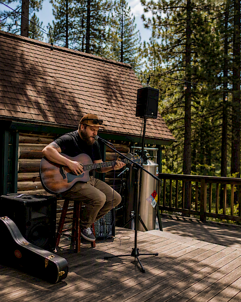 An outdoor musician plays acoustic guitar on a cabin deck with a microphone, speakers, and forest trees in the background, enjoying live sound.