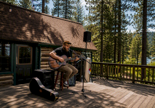 An outdoor musician plays acoustic guitar on a cabin deck with a microphone, speakers, and forest trees in the background, enjoying live sound.