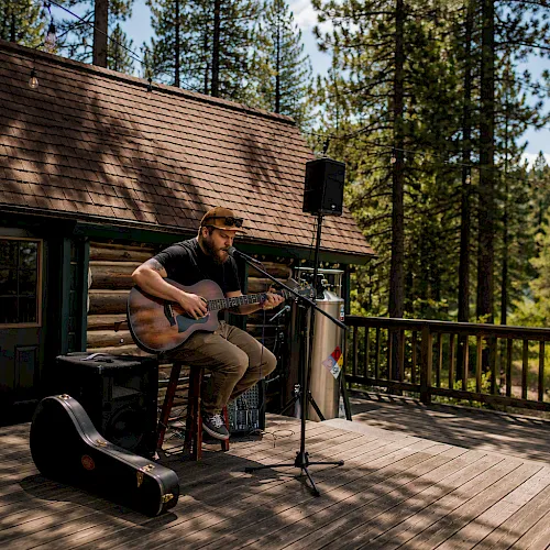 An outdoor musician plays acoustic guitar on a cabin deck with a microphone, speakers, and forest trees in the background, enjoying live sound.