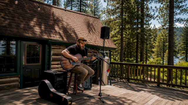 An outdoor musician plays acoustic guitar on a cabin deck with a microphone, speakers, and forest trees in the background, enjoying live sound.