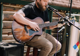 A bearded man wearing sunglasses plays an acoustic guitar on an outdoor stage into a microphone.