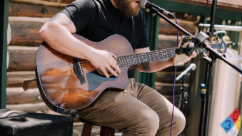 A bearded man wearing sunglasses plays an acoustic guitar on an outdoor stage into a microphone.